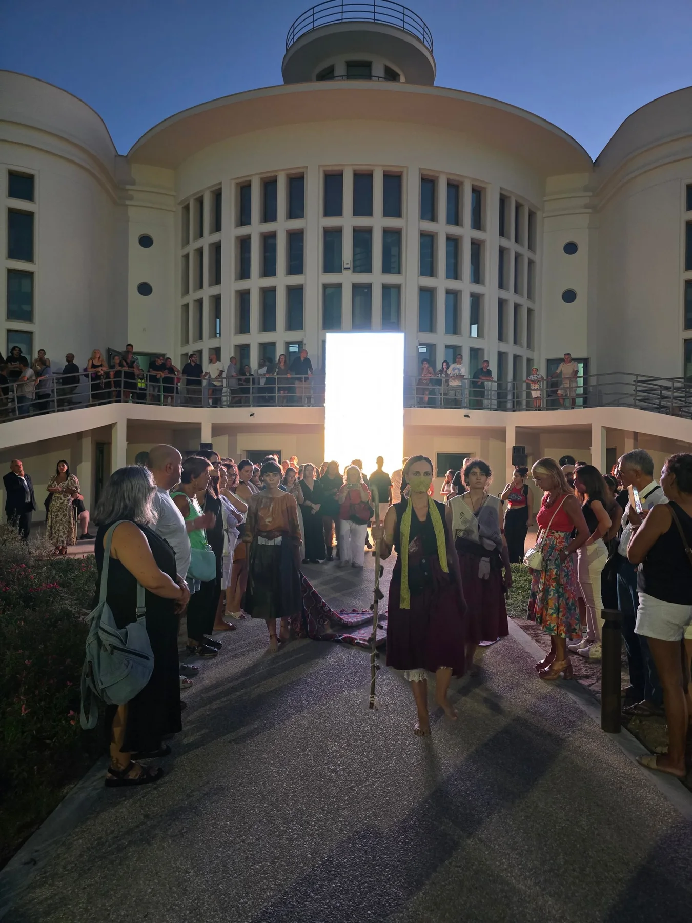Tre performer in processione davanti alla Colonia Stella Maris, con il pubblico ai lati e una porta di luce sullo sfondo