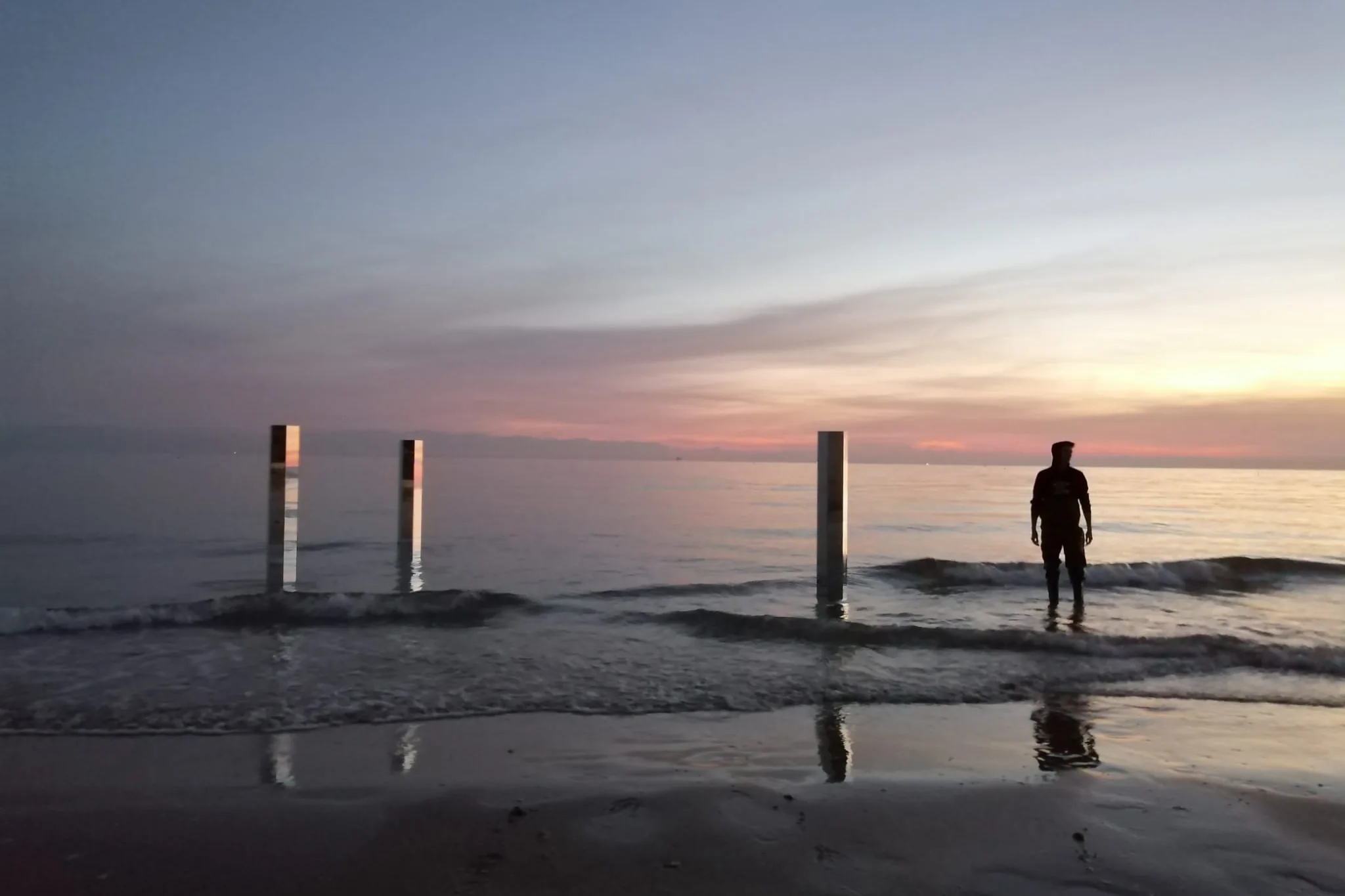 Al tramonto, colonne di specchio piantate sulla battigia e una figura in controluce nell'acqua