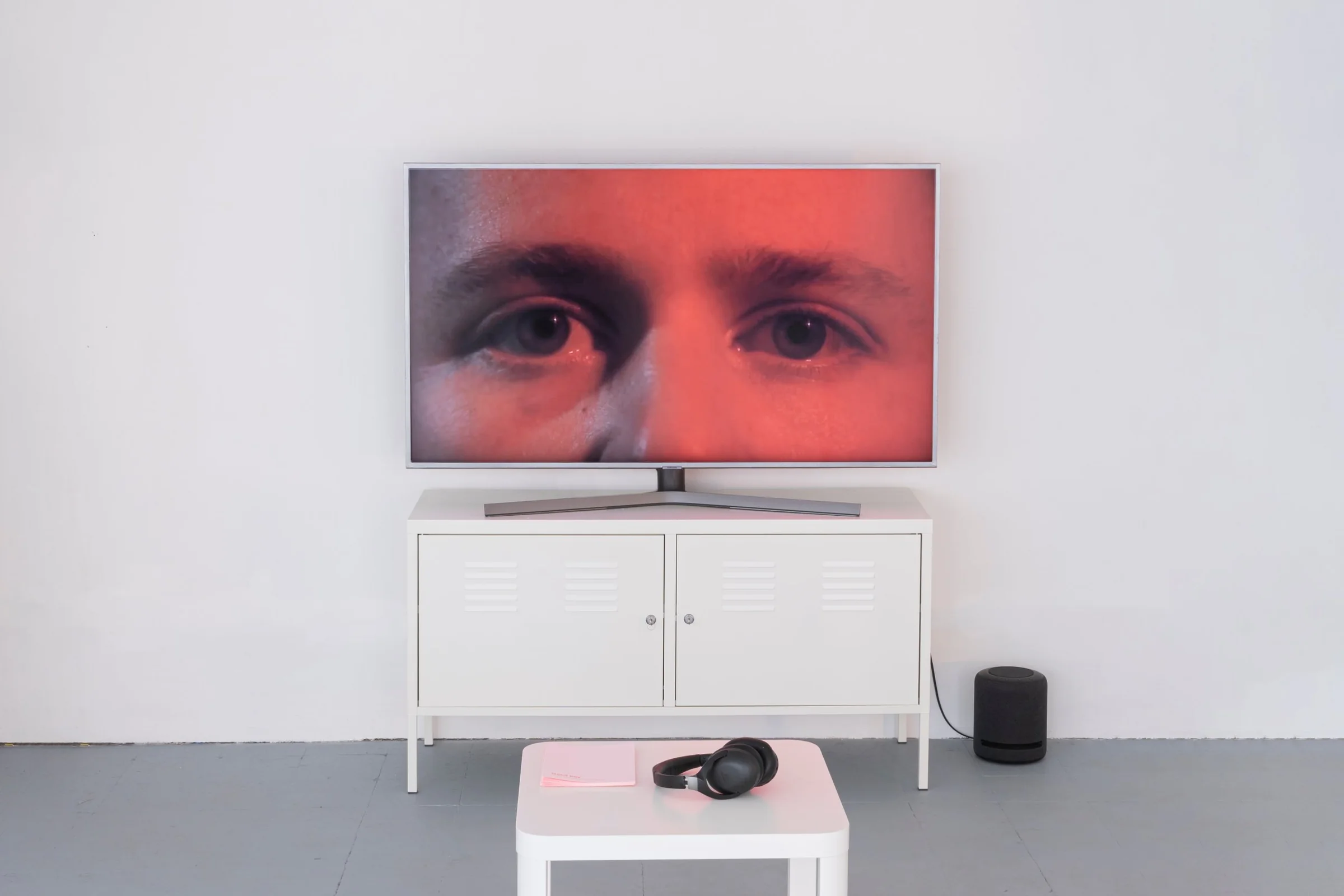 Un televisore su un mobile bianco con il primo piano di due occhi in luce rossa; davanti, un tavolino con le cuffie