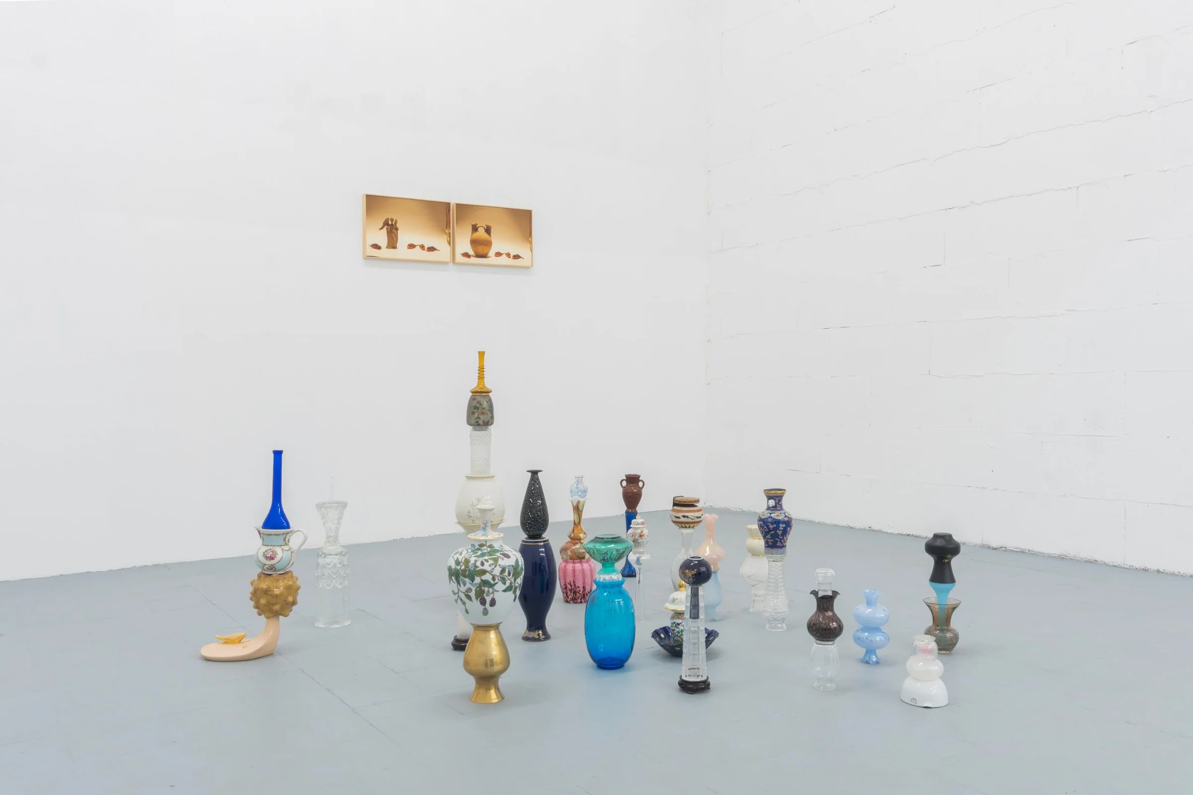 Vasi e bottiglie di vetro e ceramica sovrapposti sul pavimento grigio dello studio, due fotografie sulla parete