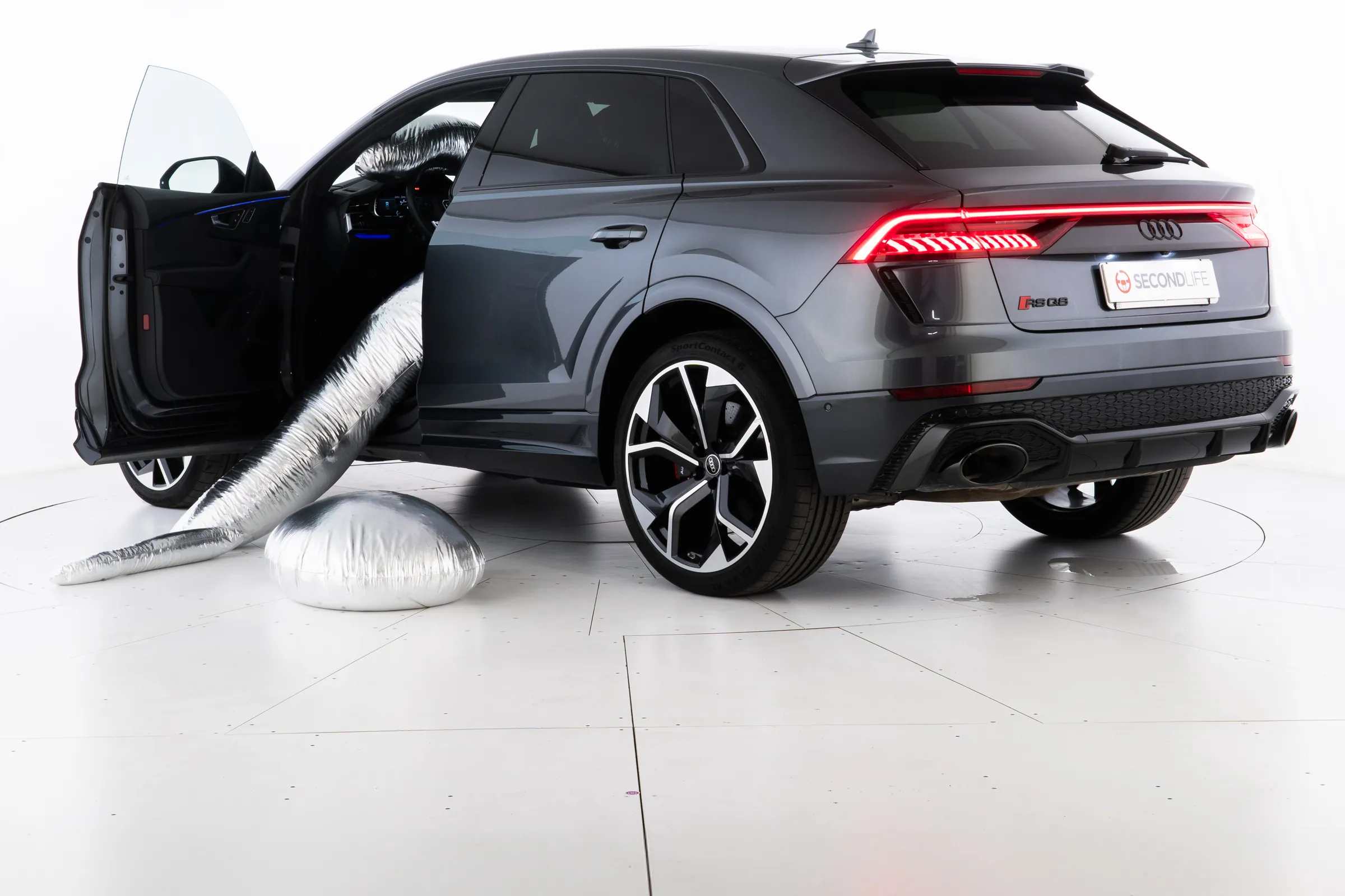 Un'auto grigia con lo sportello aperto e una scultura argentata morbida che ne esce fino a terra